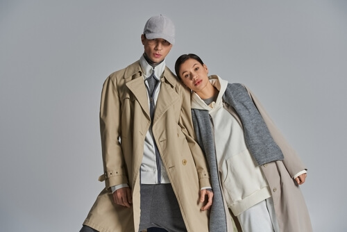Ein junges, stilvolles Paar steht souverän nebeneinander in Trenchcoats vor grauer Kulisse.